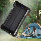 A couple sits outside a tent in a green, wooded area, holding electronic devices. A large solar power bank is prominently featured in the foreground, suggesting they are charging their devices while camping. Camping gear is visible near the tent.