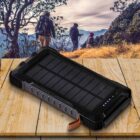 A rugged solar power bank rests on a wooden surface. Behind it, three hikers explore a scenic mountain landscape, with trees and sunlit hills in the background.