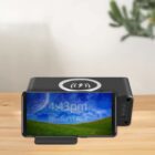 A smartphone sits in front of a black rectangular wireless charger on a wooden surface. The phone screen shows the time as 4:43 PM and the date as Tuesday, 11 April. A plant is partially visible in the top right corner.