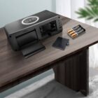 A black digital clock with a wireless charging pad on top is open, showing its battery compartment. Its placed on a wooden table, accompanied by three AA batteries and a small cover. A green plant is partially visible in the background.