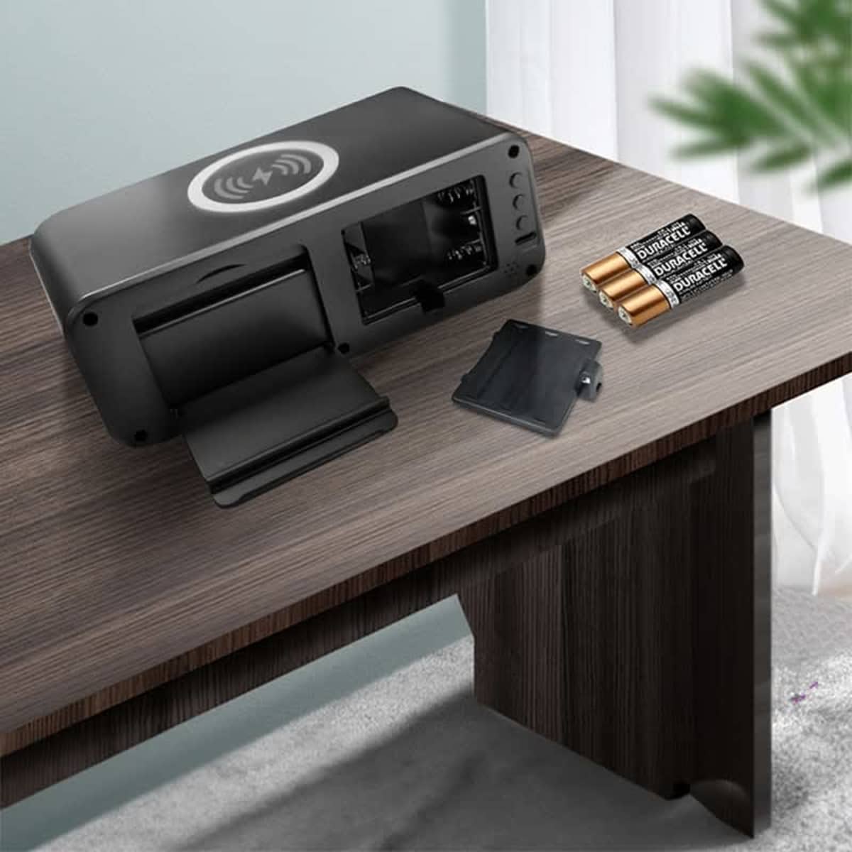 A black digital clock with a wireless charging pad on top is open, showing its battery compartment. Its placed on a wooden table, accompanied by three AA batteries and a small cover. A green plant is partially visible in the background.