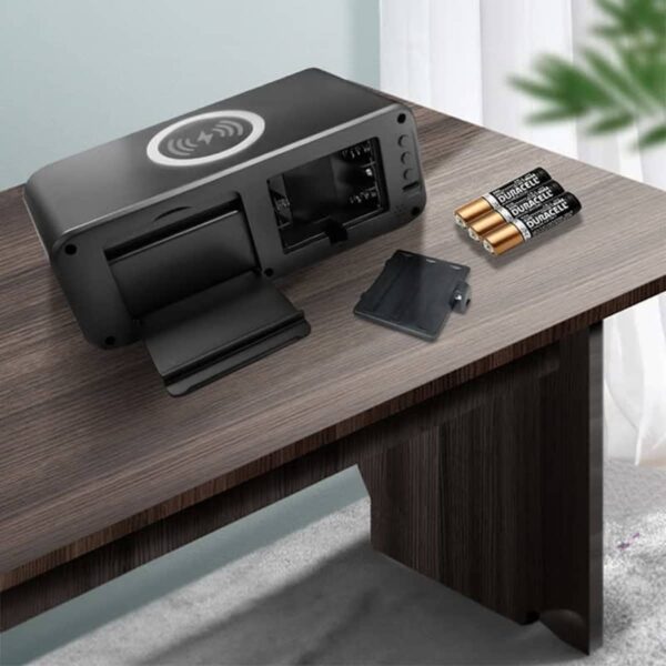 A black wireless charging alarm clock with battery slots is placed on a wooden table. Next to it are three Duracell batteries and a small black panel. A green plant is partially visible in the background.
