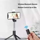 A smartphone mounted on an adjustable tripod displays a smiling woman on the screen. A hand holds a wireless Bluetooth remote control in the foreground. Text reads Wireless Bluetooth Remote and Remote Control Up to 10m.