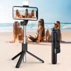 A beach scene with three women in swimwear sitting on the sand, posing for a photo. In the foreground, a smartphone on a selfie stick tripod capturing the picture. The ocean and blue sky are visible in the background.