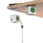 A power cube with multiple outlets and USB ports is mounted under a wooden desk. A cable is plugged into one of the outlets while a device is charging via USB. A mounting bracket secures an extension cord to the desk.