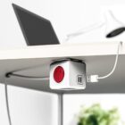 A cube-shaped power strip mounted under a white desk with multiple outlets and USB ports. A laptop and desk lamp sit on the desk, while a cable is plugged into the USB port, illustrating a tidy workspace.