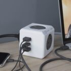 A white power cube with multiple outlets and USB ports sits on a desk, connected to various electronic devices, including a smartphone and a laptop.