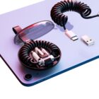 A close-up of a tablet placed on a surface. On it, there is a coiled USB-C to USB cable, with one neatly stored in a round, transparent container. A USB-A adapter lies nearby. The scene is lit with purple and pink hues.