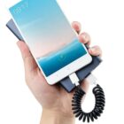 A hand holding a white smartphone connected to a portable power bank with a coiled charging cable. The phone screen displays the time 09:17 and a battery charging symbol.