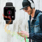 A person wearing headphones and sunglasses stands smiling while using a smartphone. A smartwatch on their wrist displays a music app. Colorful musical notes flow from the watch, suggesting music is playing. They are against a light, textured wall.