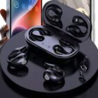 A set of black wireless earbuds with ear hooks and a charging case are displayed on a round black platform. A smartphone is visible in the background, partially held by a hand. A small treble clef symbol is also on the platform.