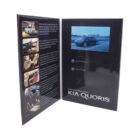 An open brochure for Kia Quoris featuring car images on the left page and a video screen displaying a car on a road on the right. The text highlights Quoris features and design.