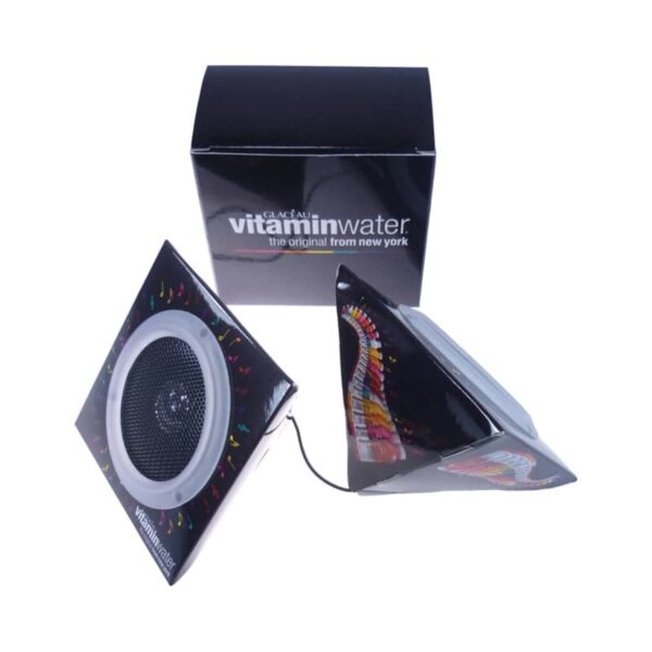 A pair of triangular black speakers with colorful patterns, connected by a cable, are placed next to a black box labeled Vitamin Water. The design incorporates a white circular speaker grille on each unit.