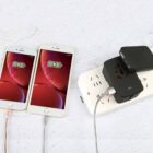 Two smartphones with charging icons displayed on screens are connected to a white power strip. The power strip has multiple outlets and a black adapter is plugged into it. The setup is on a white textured surface.