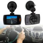 Car dashboard camera shown in two angles, front and back. The front view displays a video screen showing a road with mountains in the distance. The back view shows the lens and buttons. A person is driving a car, holding the steering wheel.