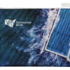 A zippered pouch featuring an aerial view of ocean waves crashing against a blue-tiled swimming pool. The words Destination NSW are printed on the upper left corner.