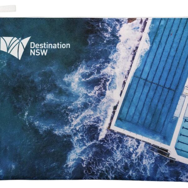 A pouch displaying an aerial view of an ocean pool with waves crashing against it. The design includes the Destination NSW logo in the top left corner. The pool is rectangular with visible lanes and surrounded by white borders.