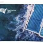 A zippered pouch featuring an aerial view of a coastal swimming pool with ocean waves crashing against it. The words Destination NSW are printed in white in the top left corner.