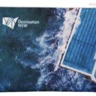 A zippered pouch featuring an aerial view of ocean waves crashing against a swimming pool. The pool is situated at the edge of the water. Destination NSW is printed on the top left corner.