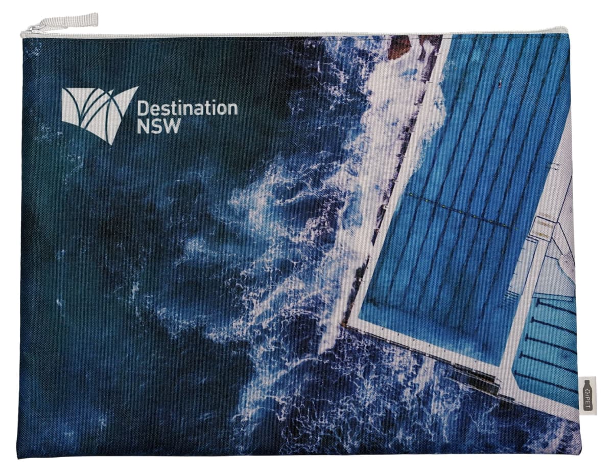 Aerial view of an ocean pool with waves crashing against it. The pool is rectangular, with clear blue water, and is situated next to the ocean. The image features the text Destination NSW in the upper left corner.