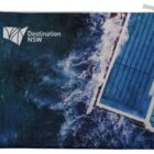 Aerial view of ocean waves crashing against a rectangular swimming pool. The pool has clear blue water and lane markings. The text Destination NSW and a logo are visible on the top left corner of the image.