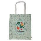 A green tote bag featuring a Farmers Market design with colorful illustrations of vegetables like corn, tomatoes, carrots, and radishes. The background is patterned with small vegetable icons. The text reads Natural Products Since 1881.