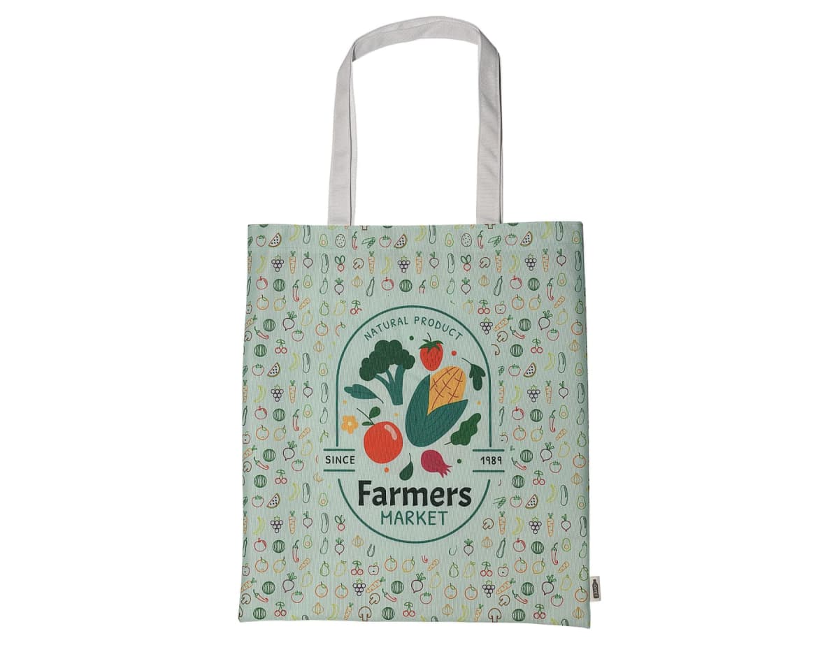 A green tote bag featuring a Farmers Market design with colorful illustrations of vegetables like corn, tomatoes, carrots, and radishes. The background is patterned with small vegetable icons. The text reads Natural Products Since 1881.