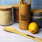 Bamboo kitchen utensils in a Nestlé holder, alongside jars of rice, lentils, and a gray container with wooden lids. A bamboo spoon with the Nestlé logo and a lemon are on a white tiled surface.