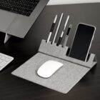 A modern workspace setup with a gray organizer featuring slots holding pens and a smartphone. A wireless mouse rests on the organizers surface, next to a laptop and a keyboard on a dark desk.
