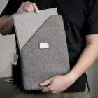 A person in a black shirt holds a gray fabric laptop sleeve with a rectangular white label in the center. The sleeves design features a darker gray section at the front. The persons head is not visible in the image.