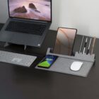 A tidy workspace featuring a laptop, wireless keyboard, and mouse. A gray organizer holds a smartphone, tablet, and pens, all neatly arranged on a dark desk surface. The laptop screen displays a serene beach scene.