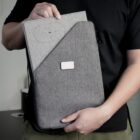 A person holds a gray laptop sleeve with a fold-over flap. The sleeve features a rectangular patch and a circular pattern on the flap. The person wears a black shirt and green pants. The background is blurred.