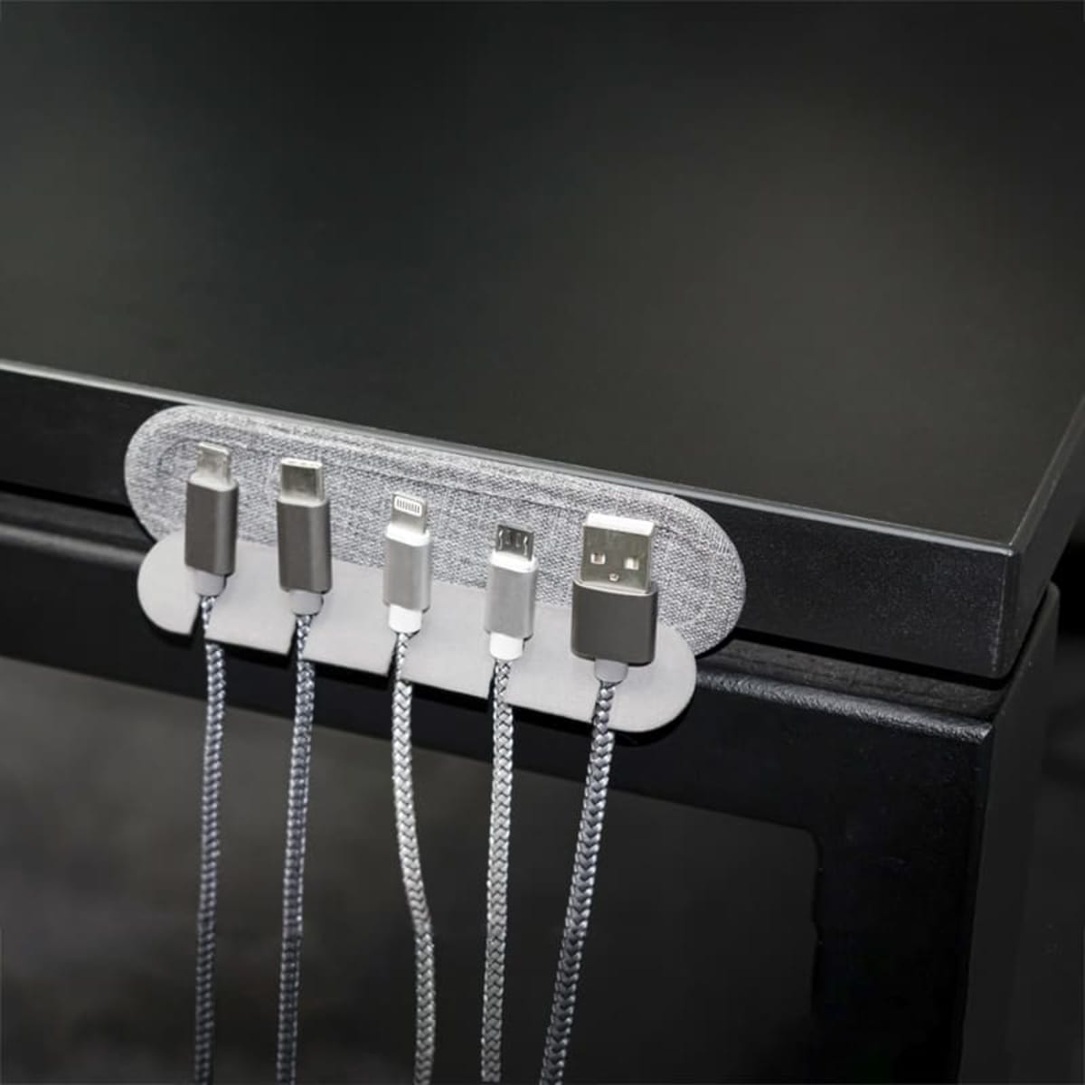 A cable organizer attached to the edge of a dark desk holds five different types of charging cables, arranged in a row for easy access. The cords are neatly secured in the organizer slots.