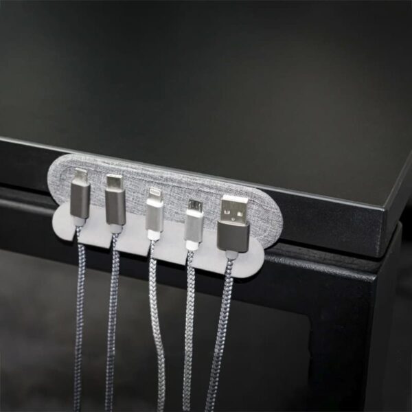 Close-up of a desk edge with a cable organizer attached. The organizer holds five different charging cables, each with a different connector type, neatly arranged side-by-side. The desk and organizer have a sleek, modern design.