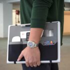 Person wearing a green sweater holds a gray multi-pocket organizer. It contains a laptop, smartwatch, smartphone, cards, and a pen. A silver wristwatch is visible on the persons left wrist. Background is a blurred indoor setting.