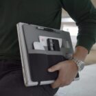 A person in a green shirt holding a laptop with an attached organizer. The organizer holds a phone, power bank, pen, earphones, and small notebook. The person is wearing a silver wristwatch. The background is blurred.