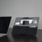 A laptop is open on a desk. Next to it, a black and gray desk organizer holds a smartphone, a white wireless mouse, a pen, and a design-related book or magazine. A card is partially visible in one of the pockets.