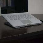 A silver laptop is open and propped up on a black desk stand. The screen is black and the keyboard is visible. The desk is dark-colored, and the background shows a blurred window.