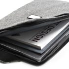A gray fabric laptop sleeve is partially open, revealing a laptop and a book titled DESIGN inside. The sleeve has a black zipper and a small, rectangular label on the front.