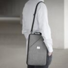 A person wearing a light-colored shirt and dark pants is seen from behind, carrying a gray laptop bag with a shoulder strap. The setting appears to be an indoor, minimalist space with neutral tones.