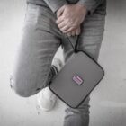 A person wearing gray jeans and white sneakers holds a gray pouch with a black zipper and a pink label that reads Rover. The person is leaning against a white surface.