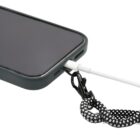 A smartphone in a dark case with a charging cable plugged into the bottom. Attached to the phone is a black and white patterned lanyard connected to a metal clip for easy carrying.