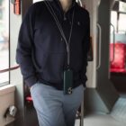 A person wearing a dark blue hoodie and light blue pants stands inside a bus, with hands in pockets. A phone hangs from a neck strap. Red seats are visible in the background.