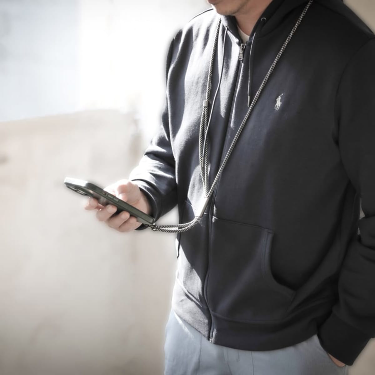 A person wearing a dark hoodie with a chain attached, holding a smartphone in one hand and standing against a bright, blurred background. The light casts soft shadows, emphasizing the casual attire and relaxed posture.
