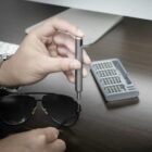 A person is using a small screwdriver to tighten the hinge of black sunglasses. A screwdriver set with various bits is visible on a dark wooden table beside a keyboard and a partially visible screen.