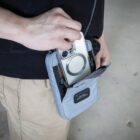 A person is placing a smartphone with a visible circular magnetic ring into a light gray fabric pouch worn around their neck. The pouch has a flap and a small label. They are wearing beige pants and a black top.