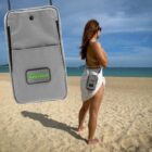 A person stands on a sandy beach wearing a white outfit, with their back to the camera. Theyre holding a small gray bag over their shoulder. The sky is clear and the ocean is calm. A close-up image of the bag with the logo SOLO ECO is superimposed.