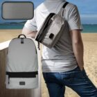 A person in a gray t-shirt and jeans stands on a sandy beach with water in the background, wearing a small gray crossbody bag. A larger matching gray backpack is displayed in the foreground, with a small close-up insert of the materials texture.