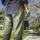 A person wearing light green pants and a dark shirt holds a smartphone with a strap in their hand while walking on a park path. Trees and a gazebo are visible in the background.