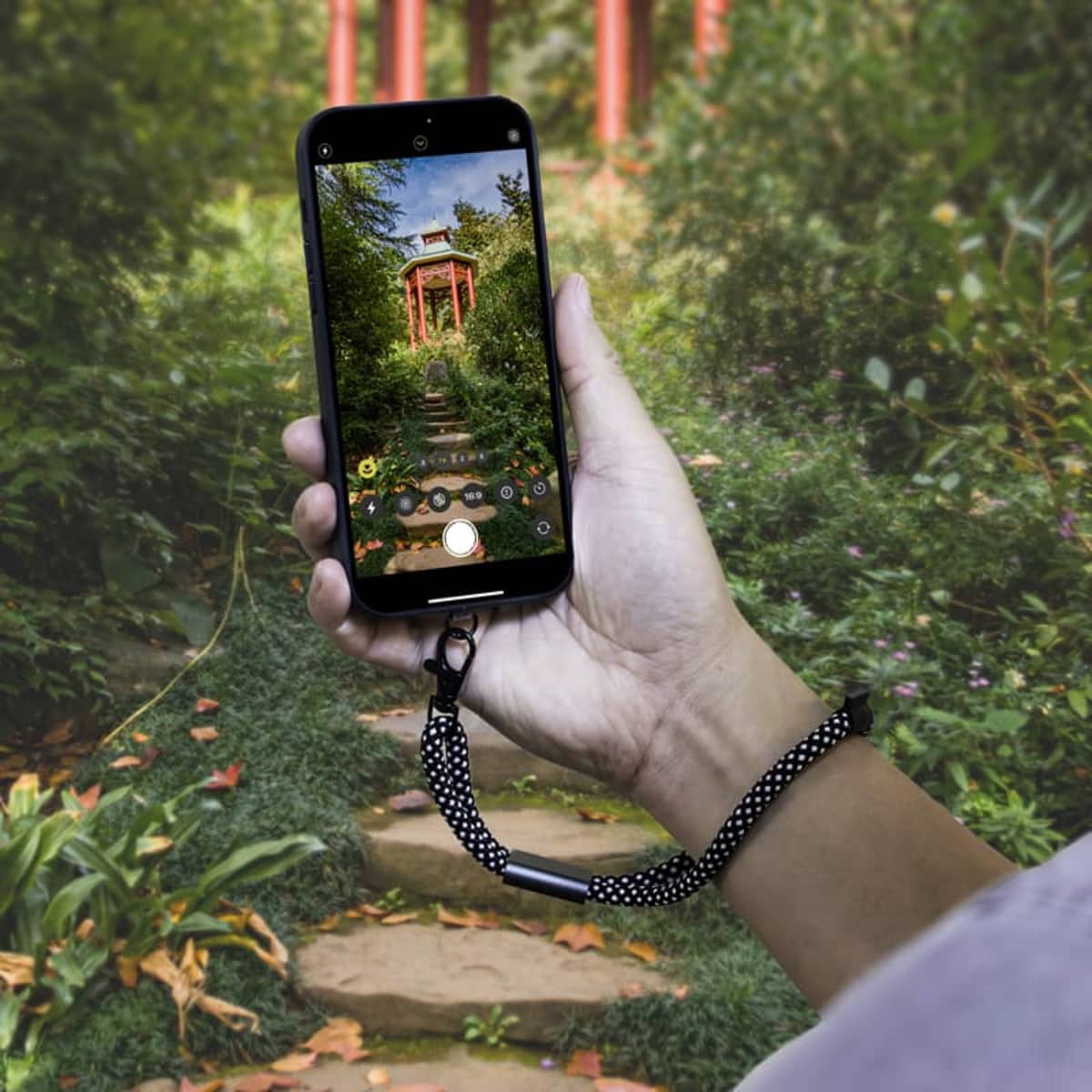 A hand holding a smartphone with a polka dot strap, capturing a photo of a garden pathway leading to a small, red pavilion surrounded by lush greenery. The phone screen displays the same scene.